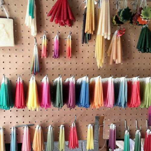 A variety of colorful candles hang on a pegboard wall, organized by size and color. The display, much like the meticulous arrangement of Gneiss Spice's Beeswax Birthday Candles, includes tapers and uniquely shaped candles, creating a vibrant and diverse collection.